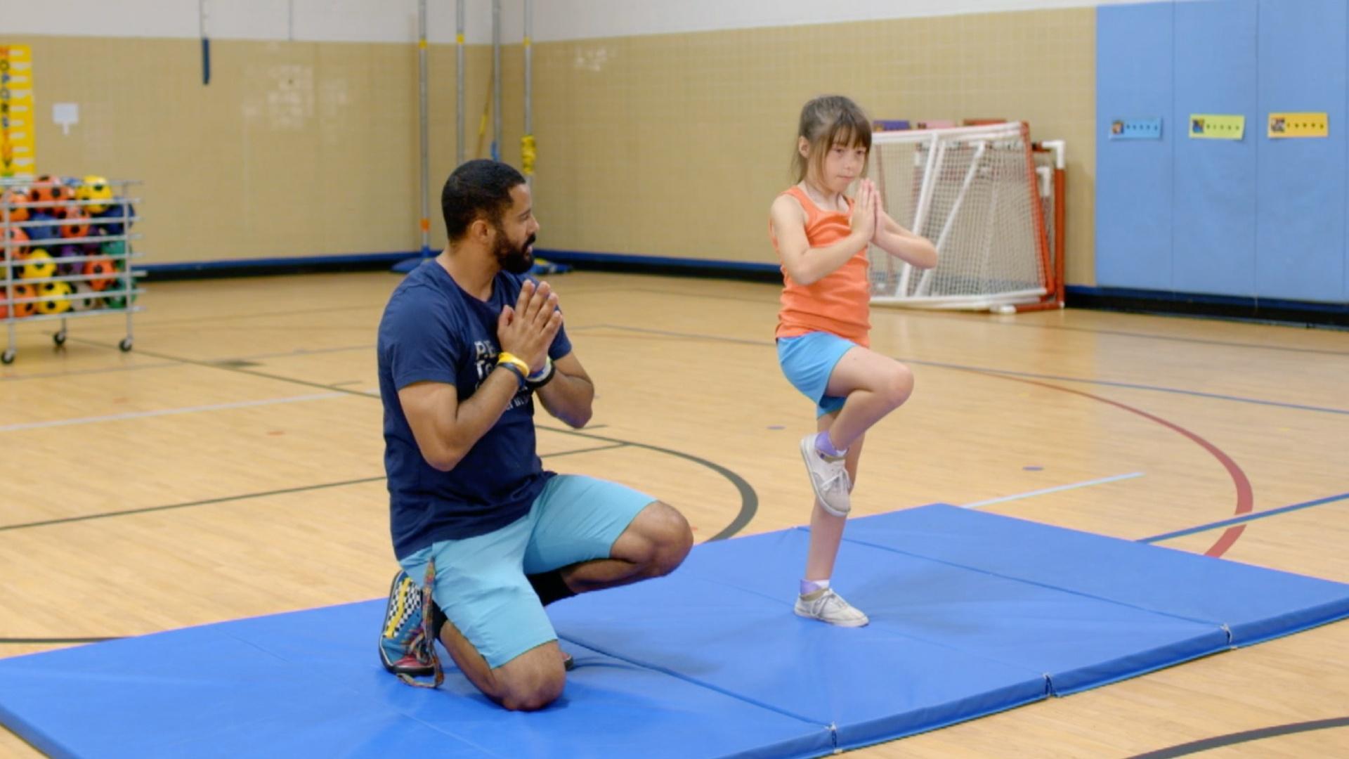 PE teacher David helps Kelsey with balance poses. Kelsey learns what a gym teacher does.